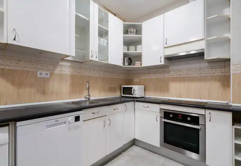 Cocina compacta y moderna con muebles blancos, horno y lavavajillas integrados, microondas sobre la encimera y fregadero de acero inoxidable; salpicadero de azulejos neutros y distribución ordenada, ideal para cocinar a diario.