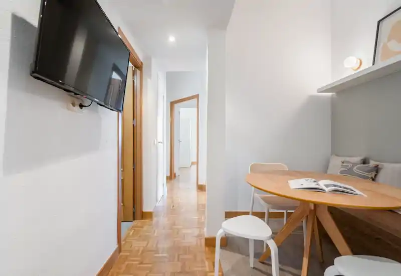 Zona de estar compacta y luminosa con mesa redonda, banco con cojines y televisor en pared; tonos neutros y suelo de parquet crean un ambiente moderno y acogedor.