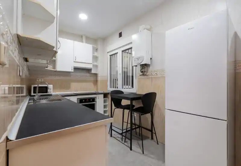 Cocina compacta y moderna con encimeras negras, horno integrado, frigorífico alto y una pequeña mesa alta para dos — luminosa y ordenada.