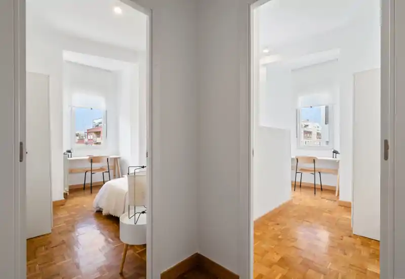 Dormitorio minimalista y luminoso visto a través de una puerta interior, con suelo de parquet, lámpara de mesita y un pequeño escritorio junto a la ventana. Paredes blancas y luz natural crean un ambiente tranquilo y funcional.