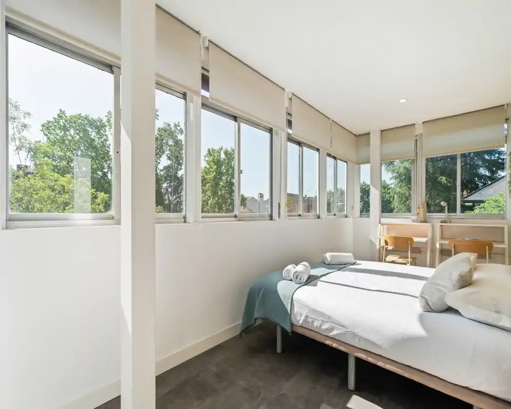 Bright modern bedroom with large windows, a neatly made double bed with pillows and a throw, and a minimalist desk area at the far end — plenty of natural light and calm neutral tones.