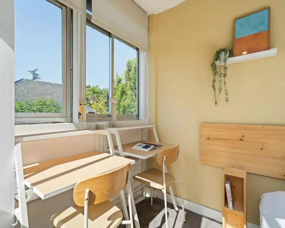 Bright, compact bedroom corner with a wooden headboard, twin desk area and large window bringing abundant natural light — ideal for a study-and-sleep setup.