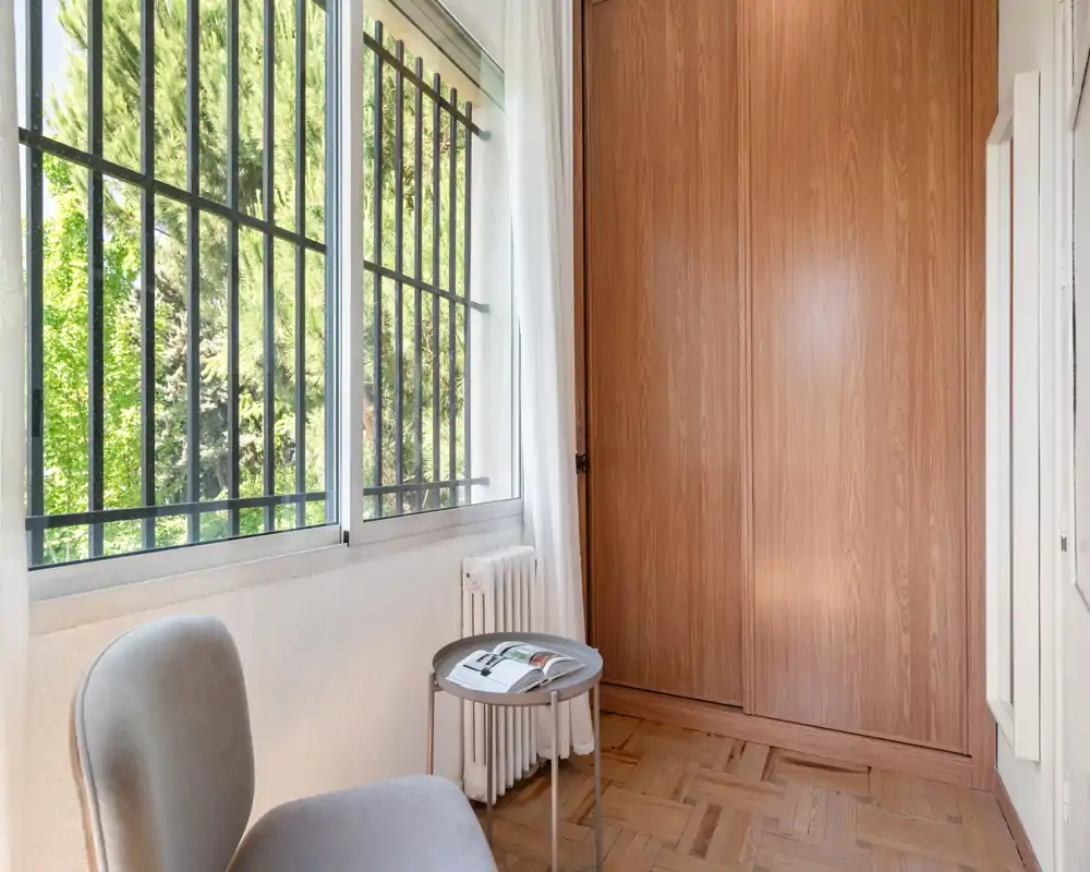 Petit coin lumineux avec fauteuil, table d'appoint et grand placard en bois près d'une fenêtre donnant sur la verdure — parfait comme coin lecture cosy en chambre.