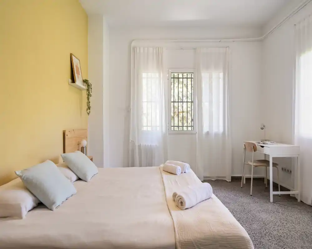 Camera da letto luminosa e ariosa con un grande letto matrimoniale, biancheria morbida, una parete accentata gialla e una scrivania vicino alla finestra — perfetta per un soggiorno rilassante.