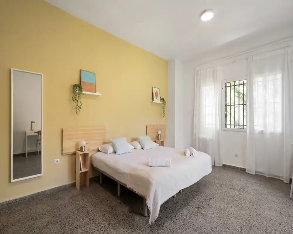Camera da letto luminosa e minimal con letto matrimoniale, biancheria chiara e luce naturale da una grande finestra; testiere in legno e parete giallo chiaro danno un'atmosfera accogliente.