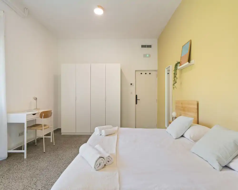 Camera da letto luminosa e minimalista con un grande letto matrimoniale, biancheria bianca, testiera in legno e asciugamani piegati. Armadio, scrivania e parete accento gialla creano un ambiente caldo e ordinato, ideale per soggiorni brevi.