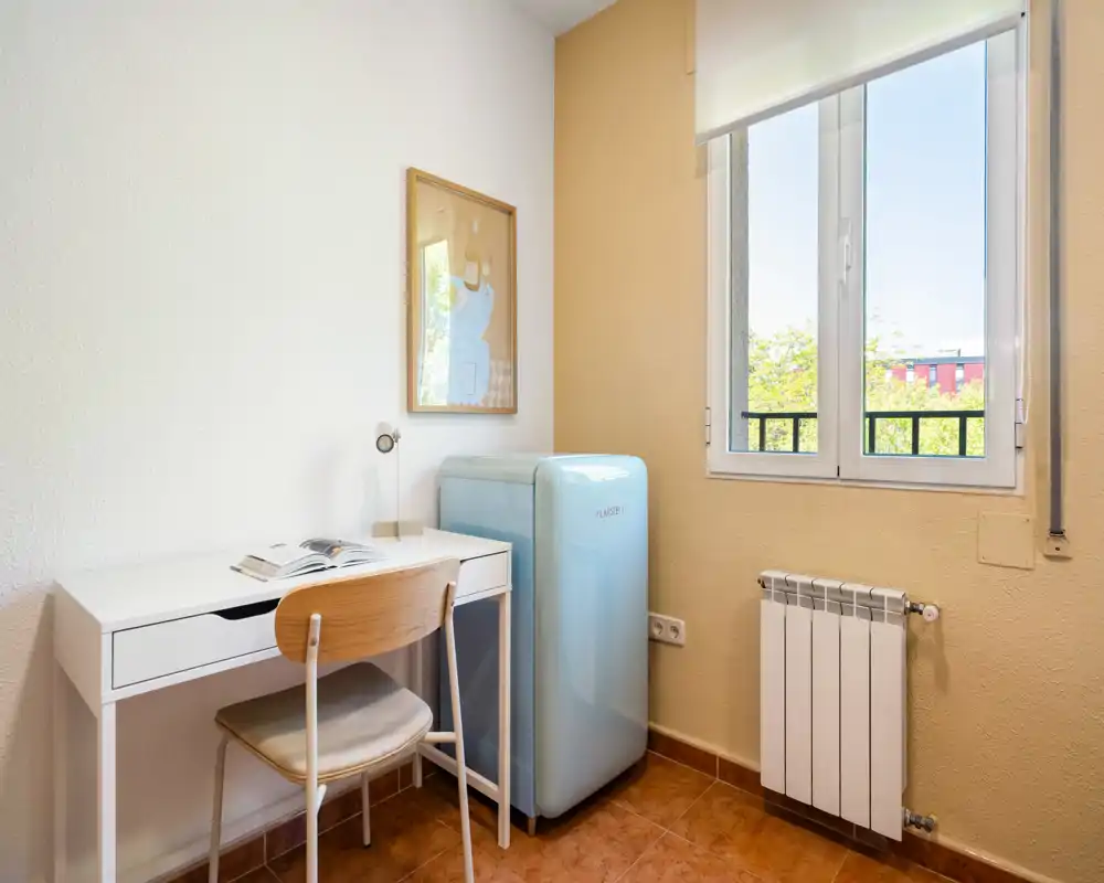 Coin cuisine compact avec un mini-frigo bleu pastel style rétro, un petit bureau blanc utilisé comme plan de travail, sol carrelé chaleureux et une fenêtre apportant de la lumière naturelle.