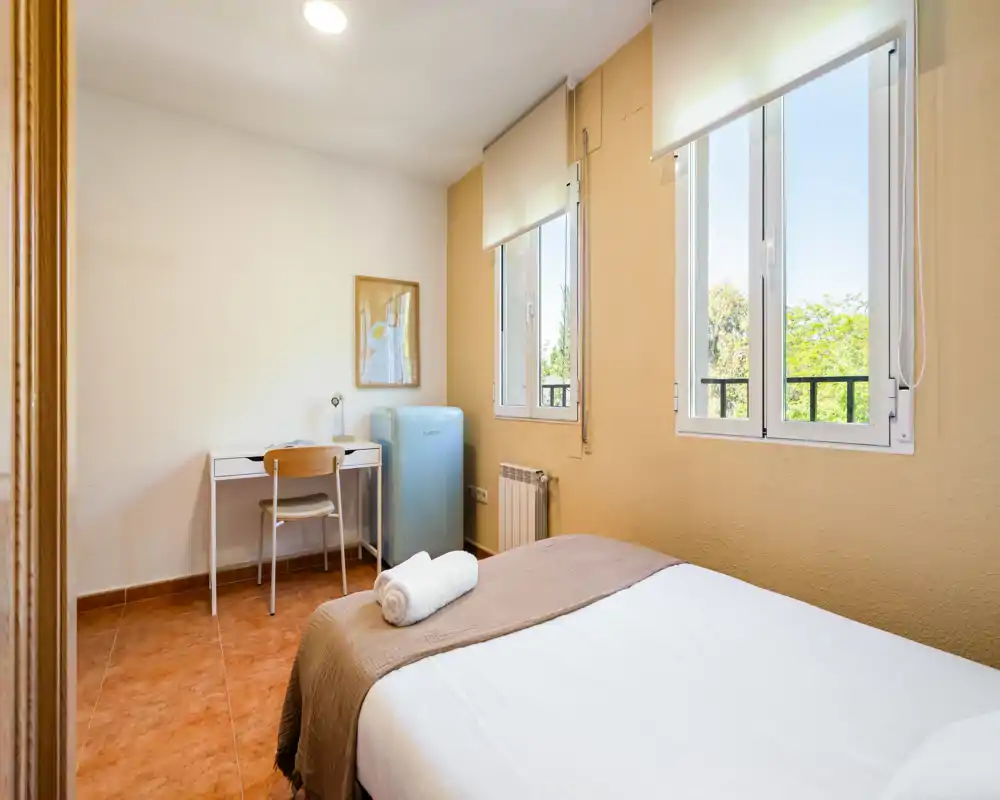 Chambre lumineuse et sobre avec lit bien fait, bureau et grandes fenêtres laissant entrer la lumière naturelle — parfaite pour une location courte durée.