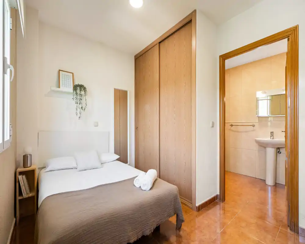 Chambre compacte et lumineuse avec un lit double, une étagère de chevet, une armoire intégrée et une salle de bain attenante visible par la porte. Décor neutre et sol chaleureux pour une atmosphère accueillante.