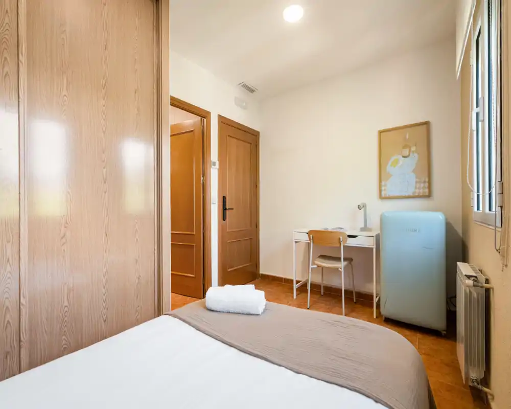 Chambre lumineuse et bien aménagée avec lit propre au premier plan, petit bureau et mini-réfrigérateur pastel — idéale pour voyageurs seuls ou étudiants.