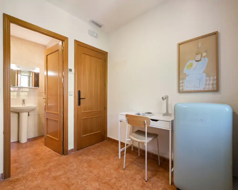 Petit hall d'entrée ou espace de passage avec un bureau compact, une chaise et un mini-frigo à droite ; des portes en bois donnent sur la salle de bains. Tons neutres et sol carrelé propre pour un espace de transition fonctionnel.