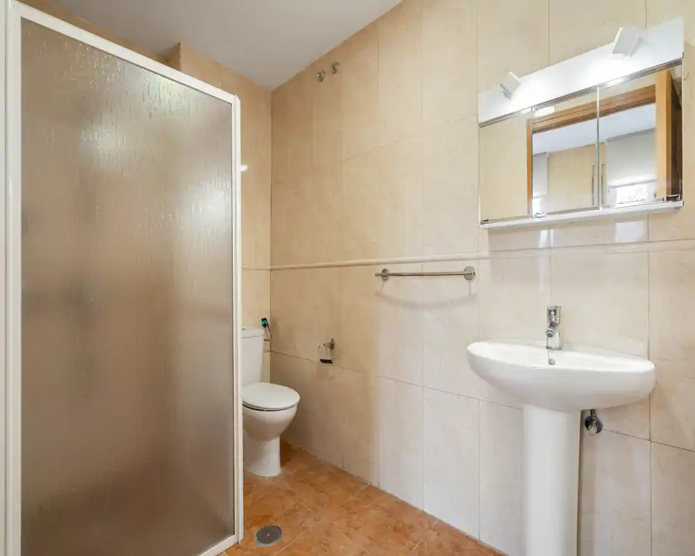 Salle de bains propre et bien éclairée avec cabine de douche, lavabo sur colonne et WC ; carrelage beige neutre et armoire-miroir au-dessus du lavabo.