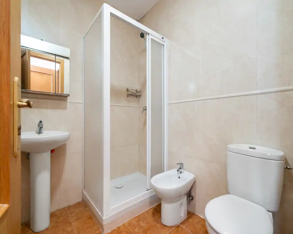 Baño limpio y compacto con cabina de ducha, lavabo pedestal, bidé e inodoro, paredes alicatadas y suelo de tonos cálidos.