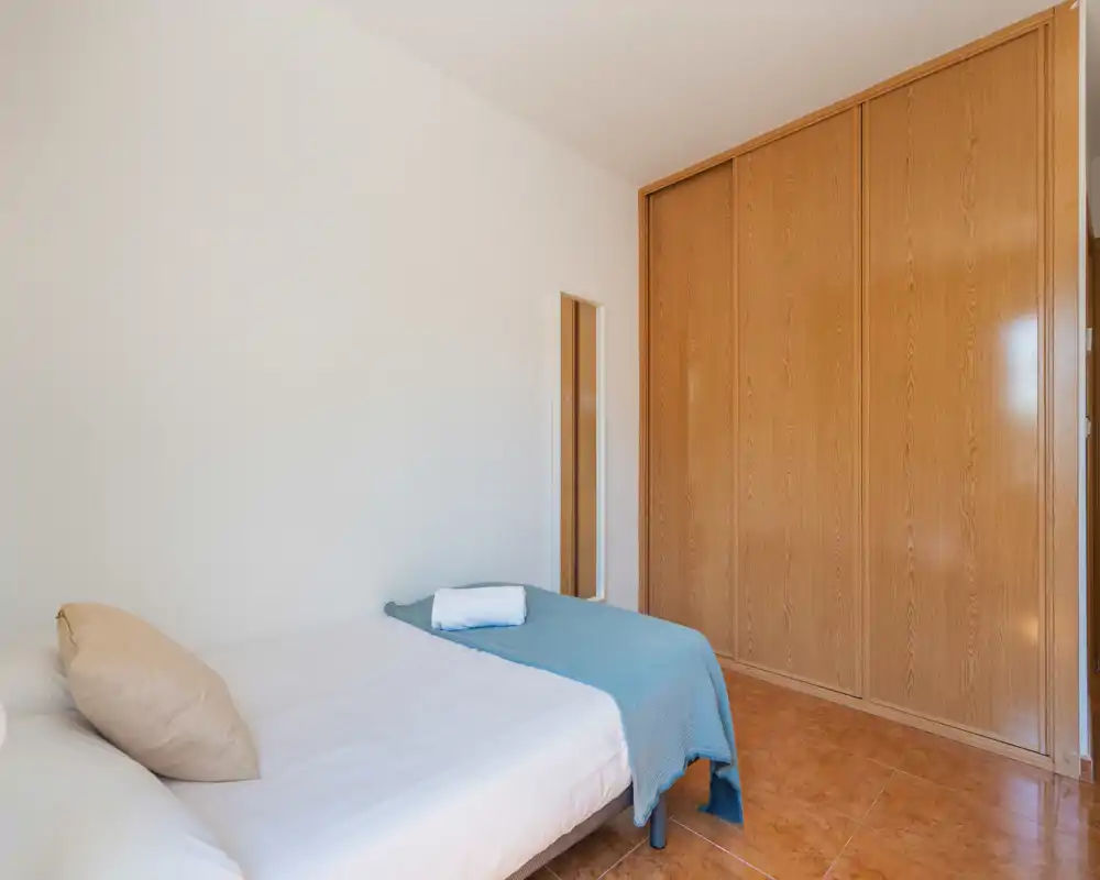 Dormitorio individual ordenado con cama hecha, armario empotrado de madera y espejo alto — luminoso, sencillo y bien iluminado.