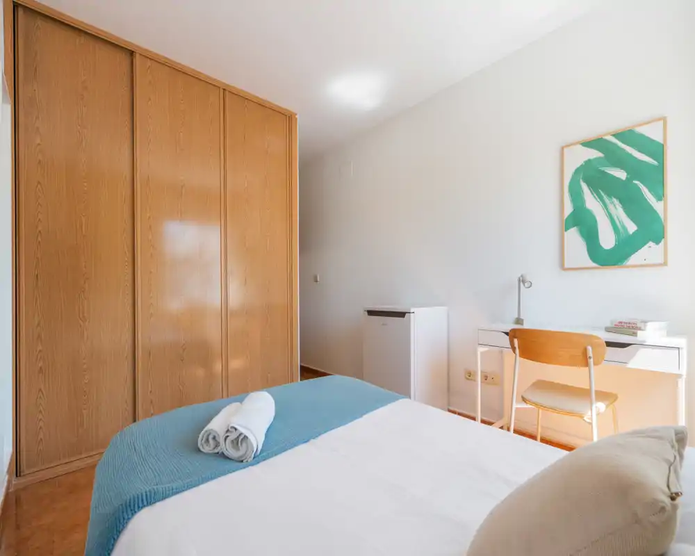 Dormitorio luminoso y sencillo con cama doble vestida en blanco y plaid azul, armario empotrado de madera y pequeño escritorio con silla: despejado y acogedor, perfecto para estancias cortas.