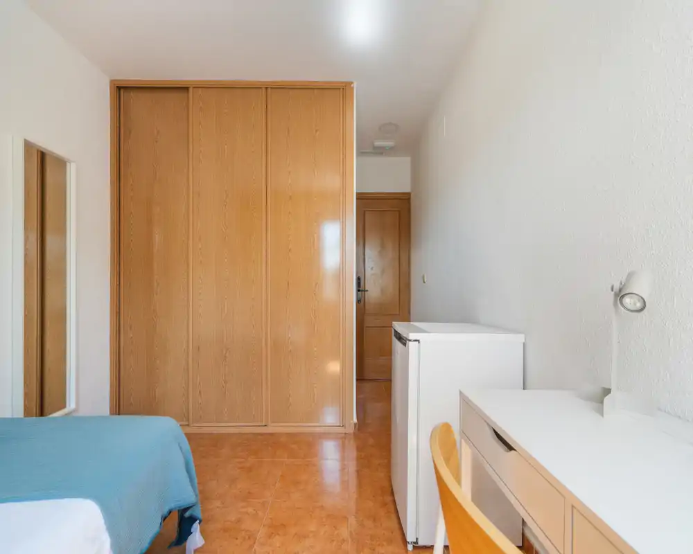 Dormitorio luminoso y sencillo con esquina de la cama visible, armario empotrado de madera y escritorio con silla y lámpara — ideal para una persona.