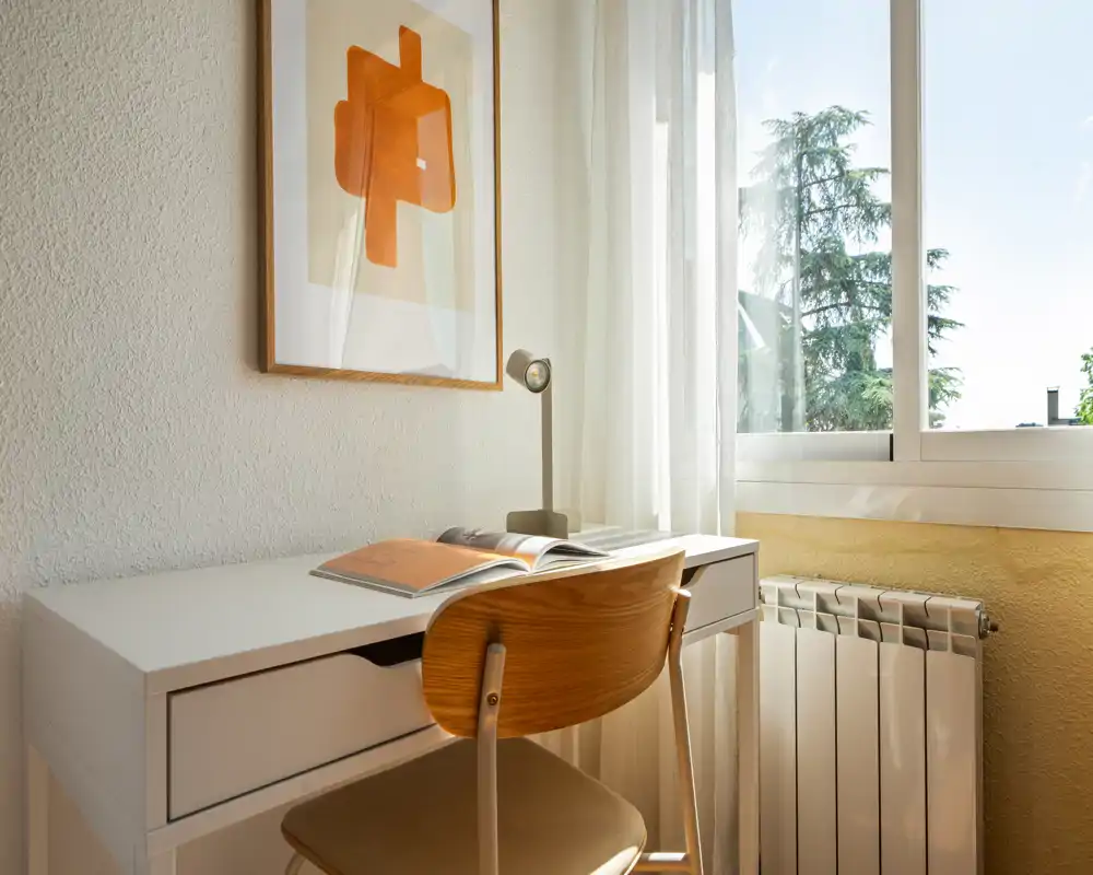 Rincón de trabajo luminoso y ordenado junto a la ventana con escritorio blanco, silla de madera, lámpara de mesa y cuadro enmarcado — ideal para estudiar o trabajar desde casa.
