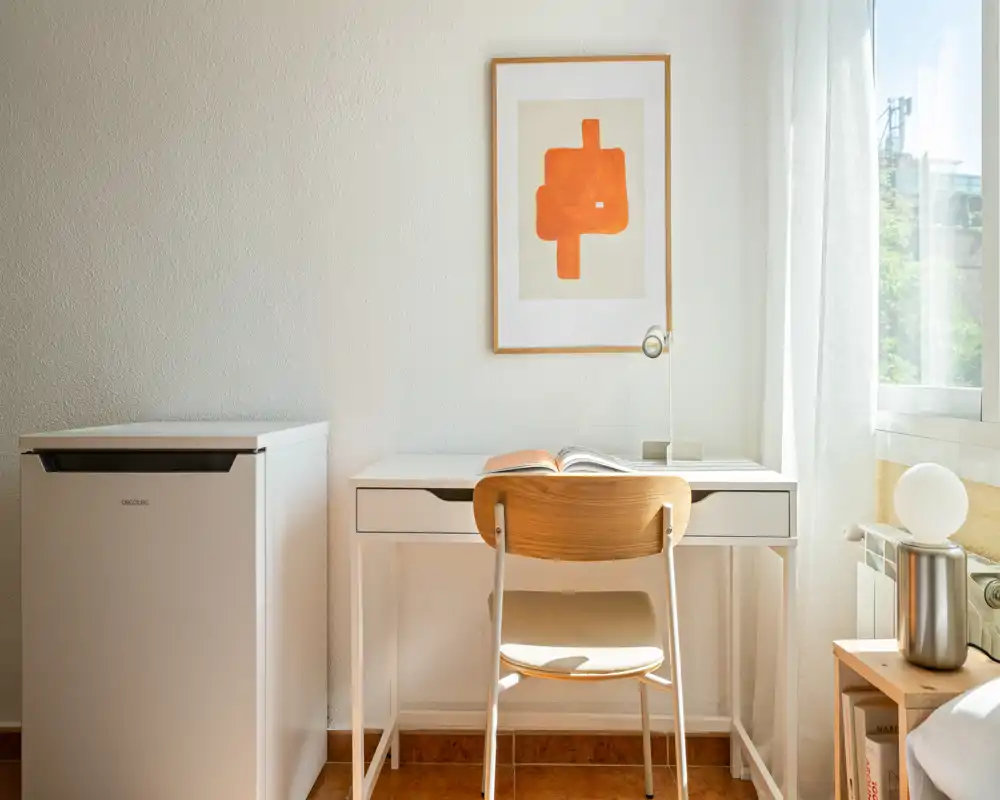 Rincón luminoso y minimalista de dormitorio con escritorio blanco, silla de madera y suave luz natural — ideal como zona de trabajo compacta.