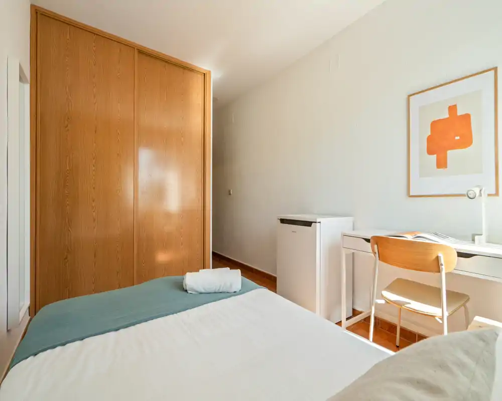 Dormitorio luminoso y sencillo con cama bien hecha, amplio armario empotrado de madera y escritorio con silla junto a la ventana — ideal para una estancia cómoda.