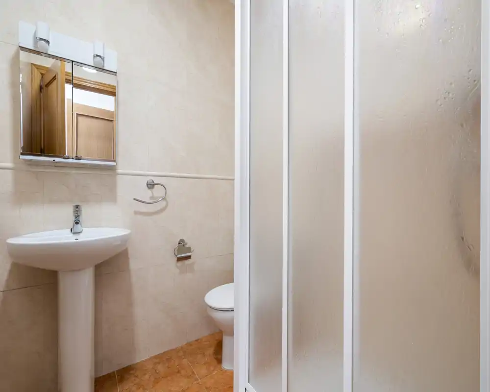 Baño compacto y luminoso con lavabo de pedestal, armario con espejo, inodoro y mampara de ducha de vidrio esmerilado.