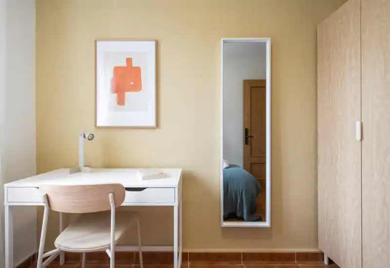Minimal, well-lit bedroom corner with a desk and chair; a vertical mirror on the wall reflects part of the bed, and a light-wood wardrobe completes the calm, neutral interior.
