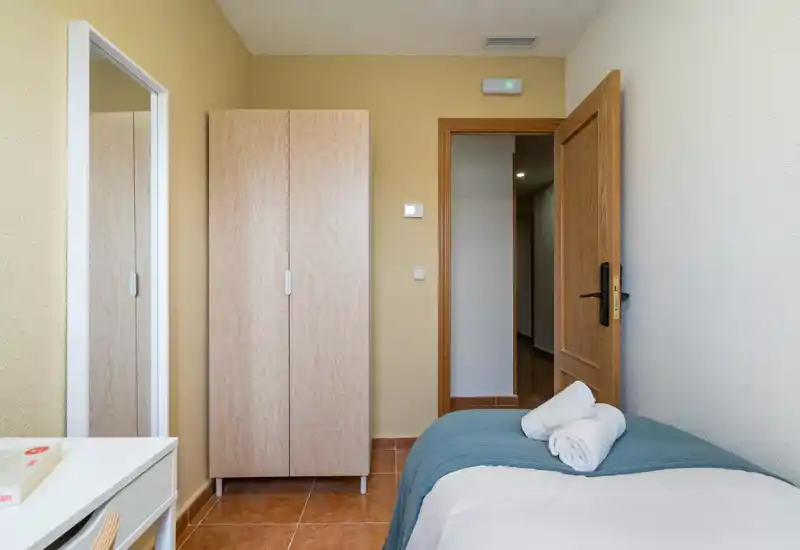 Compact, well-lit bedroom with a single bed dressed in white linens and a teal throw; wardrobe and full-length mirror maximize storage and visual space.