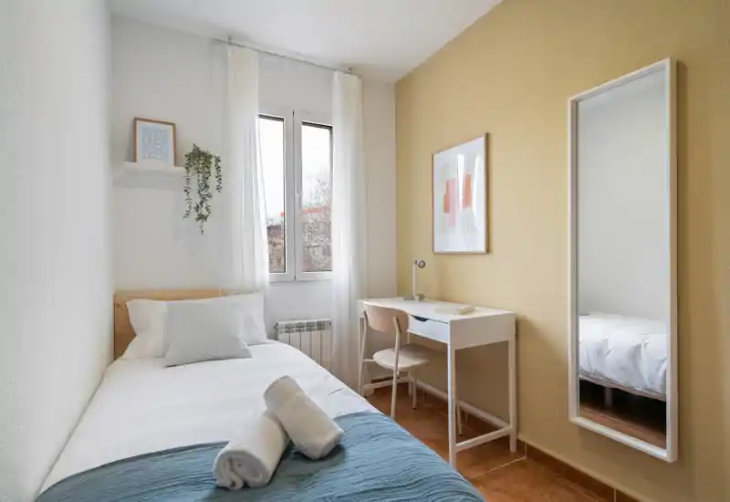 Bright, cozy single bedroom with a neatly made bed, natural light from a central window, a compact desk and full-length mirror — ideal for a solo renter.