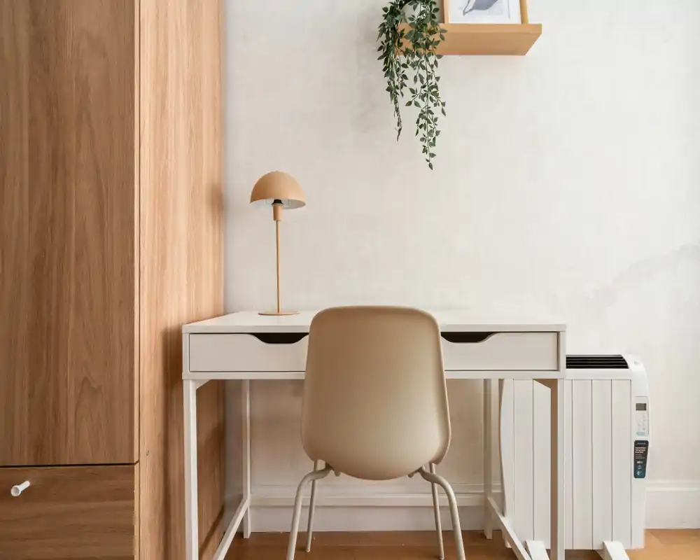A minimalist workspace corner with a white desk, beige chair, small lamp and a hanging plant against a pale wall; warm wooden cabinetry frames the left side.