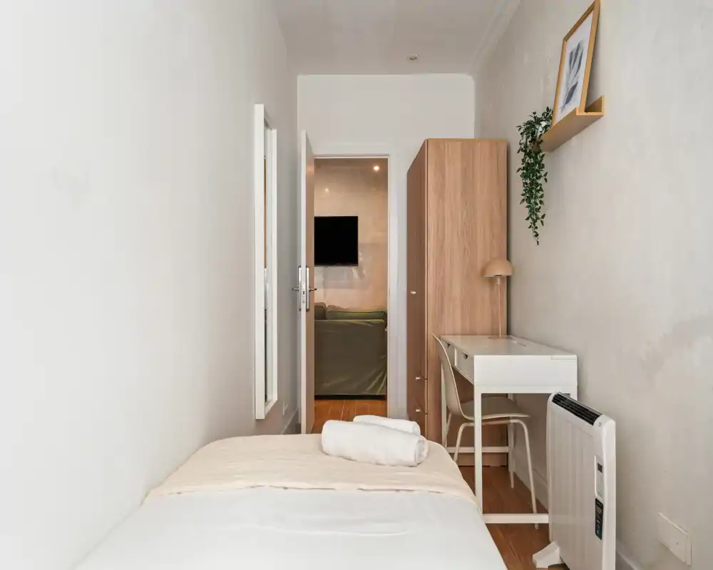 Compact bedroom with a single bed in the foreground, small desk and wardrobe along the right wall, and soft neutral decor creating a calm atmosphere.
