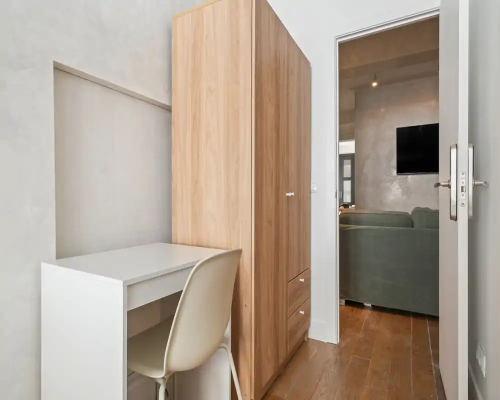 Compact bedroom corner with a white desk, chair and a tall wooden wardrobe; doorway opens onto a living area with a sofa and wall-mounted TV.