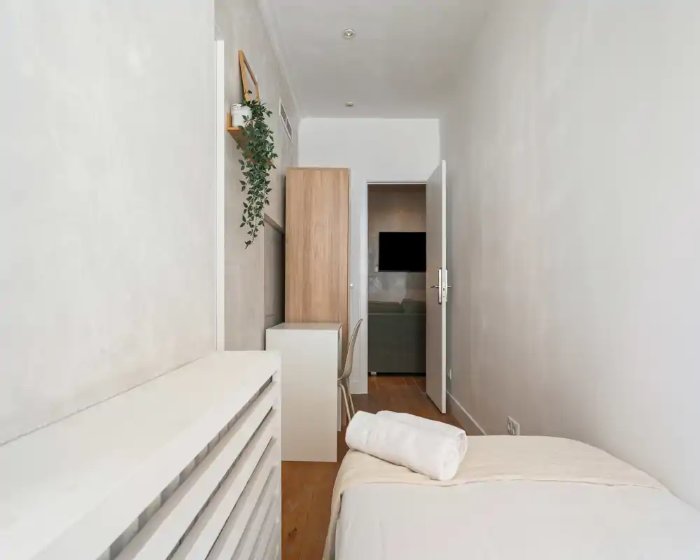 Compact minimalist bedroom with a single bed, soft neutral linens and a rolled towel; narrow layout with a small desk/wardrobe and natural, soft lighting.