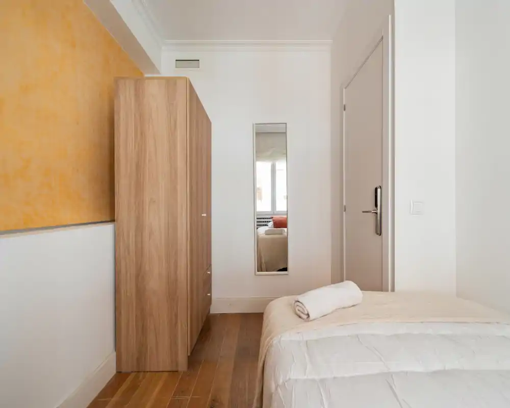 Small, simply furnished bedroom with a partially visible bed, wooden wardrobe and a tall mirror reflecting a window. Neutral tones, wooden floor and soft linens create a calm, minimal atmosphere.