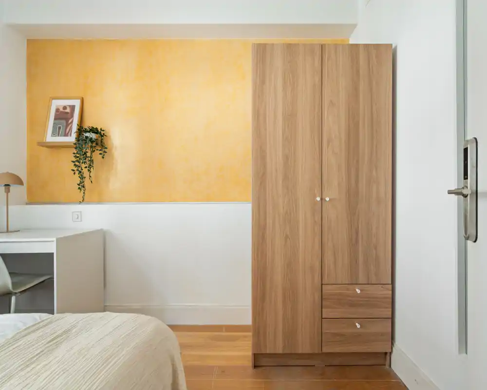 Bright, tidy bedroom corner with a visible bed edge, a wooden wardrobe and a desk under a warm yellow accent wall — calm, well-staged space ideal for rental photos.