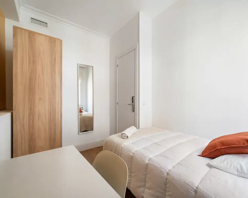 Bright, minimalist bedroom with a neatly made single bed, white duvet and an orange accent pillow. The room features a wooden wardrobe, full-length mirror and a simple desk, ideal for a single occupant or short stays.