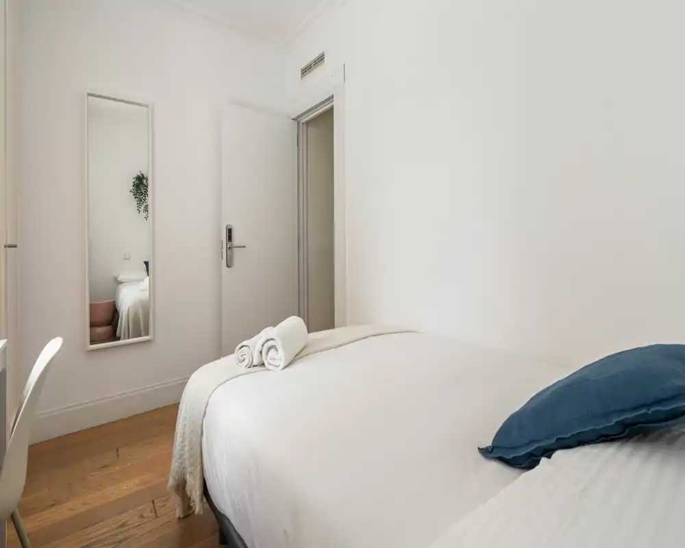 Bright, minimalist bedroom with a neatly made double bed, white linens, a blue accent pillow and rolled towels on the foot of the bed. Clean walls, wooden floor and a full-length mirror create an airy, welcoming space.