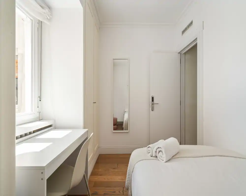 Bright, minimalist bedroom with a white bed dressed in fresh linens, a desk by the window and natural light highlighting warm wooden floors.