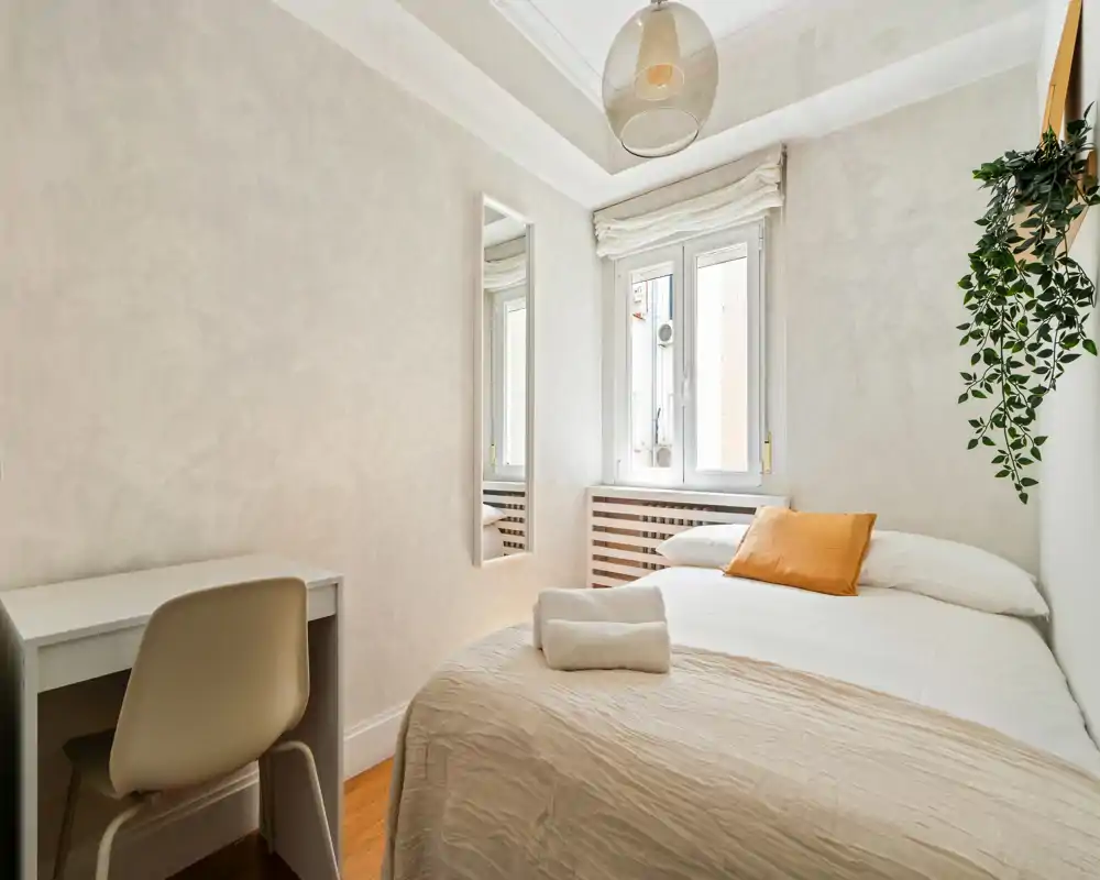 Bright, compact bedroom with a neatly made double bed, soft neutral linens and a warm accent cushion. Natural light from the window and minimal, stylish decor create a calm, welcoming atmosphere ideal for short-term rentals.