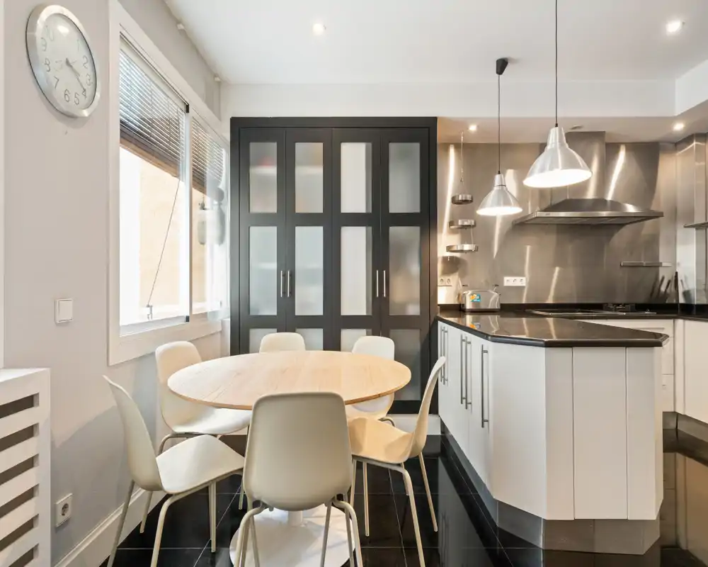 Bright modern dining area adjacent to a sleek kitchen: round wooden table with six chairs, pendant lights and built-in cabinetry, ideal for shared meals.