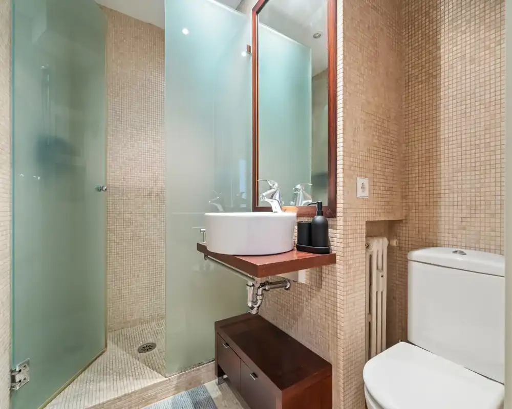 Modern compact bathroom with a walk-in shower, frosted glass panels, wall-mounted vanity with a vessel sink and a toilet; neutral mosaic tiles and warm wood accents create a contemporary look.