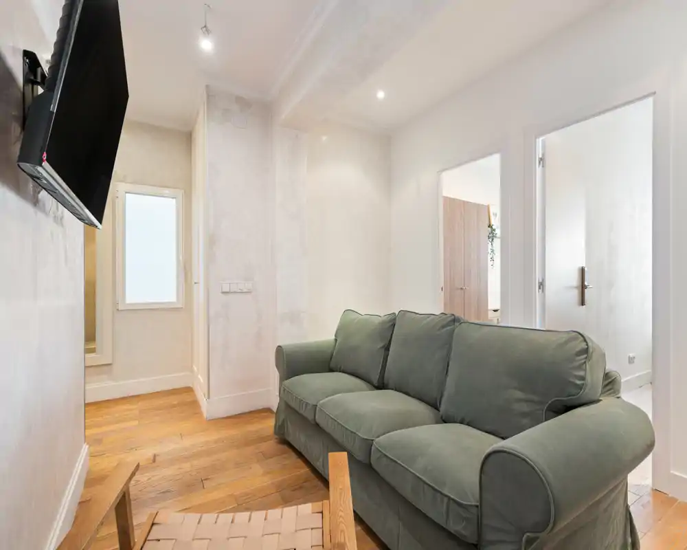 Comfortable living room with a plush three-seat sofa, wooden flooring and a wall-mounted TV. Soft neutral walls and warm natural light create a relaxed, inviting atmosphere ideal for lounging.