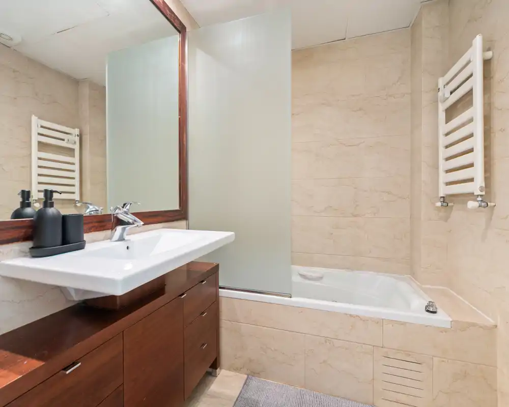 Modern tiled bathroom with a built-in bathtub, frosted glass screen, wall-mounted towel radiator and a wide sink set on wooden vanity; bright and clean.