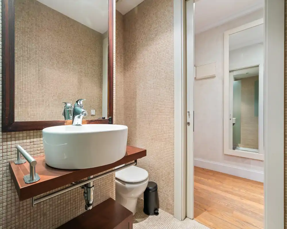 Compact modern bathroom with a vessel sink on a wooden shelf, mosaic-tiled walls, a wall-mounted mirror and a toilet. Bright, clean design with warm wood and neutral tones.