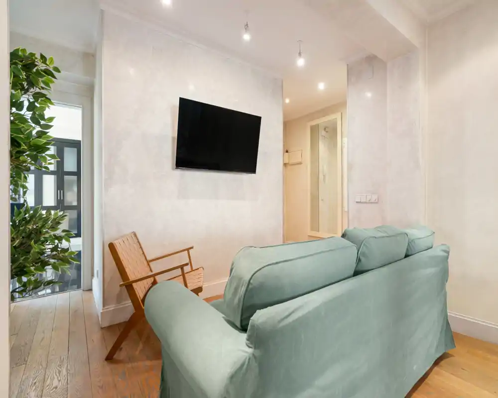 Comfortable living room with a green slipcovered sofa, wooden armchair, wall-mounted TV and warm hardwood floors — bright, well-styled space ideal for relaxing.