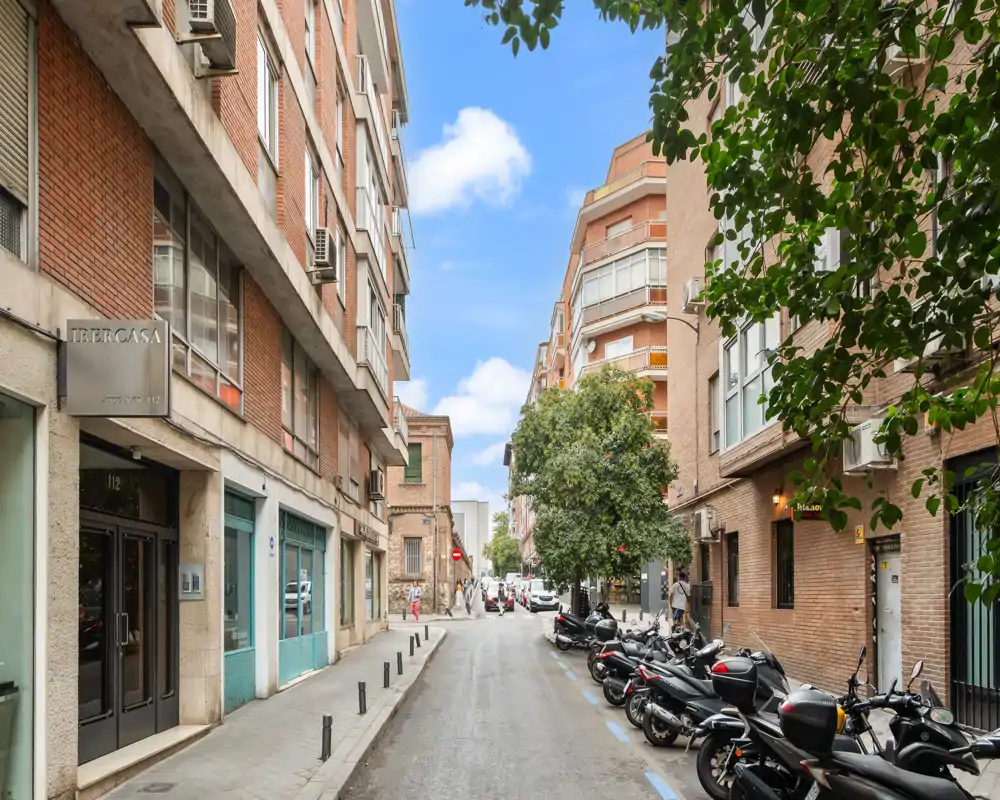 Sunny urban street scene with parked scooters, brick residential buildings, and trees lining the sidewalk — ideal to showcase a neighborhood or exterior view of an apartment.