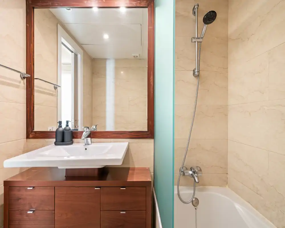 Well-lit modern bathroom with a wall-mounted sink, wooden vanity, large mirror and a bathtub with a handheld showerhead.