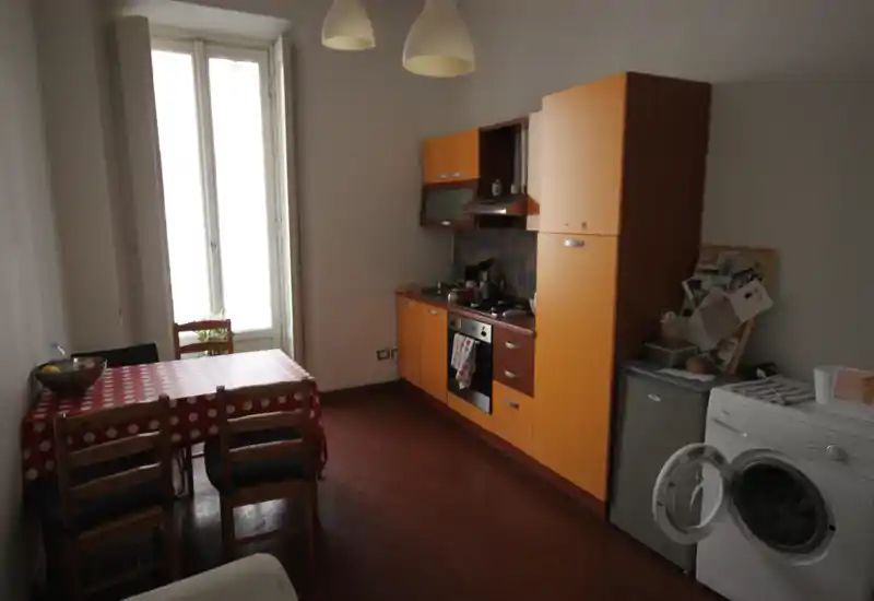 Cucina compatta con pensili arancioni, forno e lavello su una parete, tavolo da pranzo con quattro sedie vicino a una finestra alta e lavatrice in primo piano.