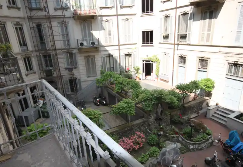 Cortile interno soleggiato visto dal balcone: area pavimentata con aiuole, cespugli e una piccola fontana/rocce, oltre a scooter parcheggiati e facciate degli appartamenti circostanti.