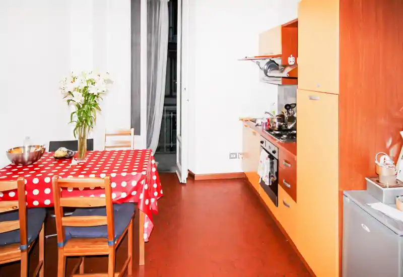 Cucina luminosa e compatta con mobili arancioni, piano cottura e un tavolo da pranzo coperto da una tovaglia rossa a pois con sedie e un vaso di fiori.