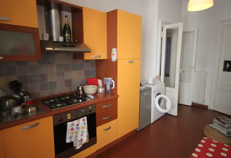 Cucina compatta e accogliente con mobili arancioni, piano cottura a gas e forno incassato; lavatrice e piccoli elettrodomestici visibili che conferiscono un aspetto pratico e vissuto.