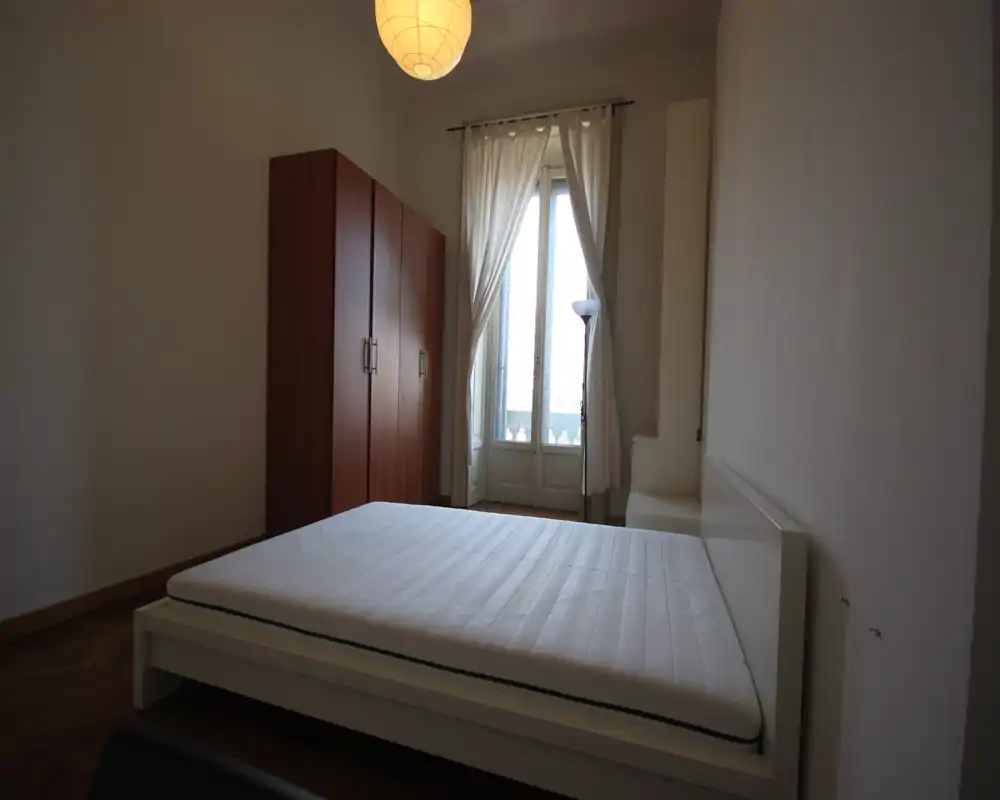 Dormitorio minimalista con cama plataforma blanca y un gran armario junto a una ventana alta con cortinas translúcidas, que proporciona buena luz natural y techos altos.